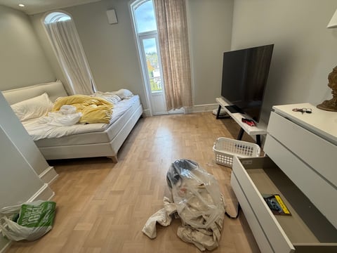 Modern bedroom with white bed, wood flooring, flat-screen TV on stand, large windows with curtains, and white furniture with moving boxes